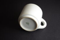 Biały, cylindryczny kubek z grubego porcelitu o szerokim uchu, z widocznymi zarysowaniami i drobnymi obiciami na krawędziach. Na spodzie naczynia znajduje się ciemnozielona sygnatura: napis „VICTORIA” z koroną powyżej oraz napis „Waffen SS” poniżej.