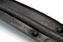 Ciężki, metalowy fragment o wydłużonym, prostokątnym kształcie, wykonany ze stali. Dłuższe boki są zagięte do góry pod kątem prostym, co tworzy kształt przypominający płytkie koryto. Pośrodku biegnie podłużne zagłębienie, a na górnej powierzchni znajdują się dwie wystające śruby. Metal jest zdeformowany i nierówny, co sugeruje działanie bardzo wysokiej temperatury lub dużego obciążenia. Na spodniej stronie widoczny jest wypukły napis z nazwą producenta Friedrich Krupp oraz fragment daty.