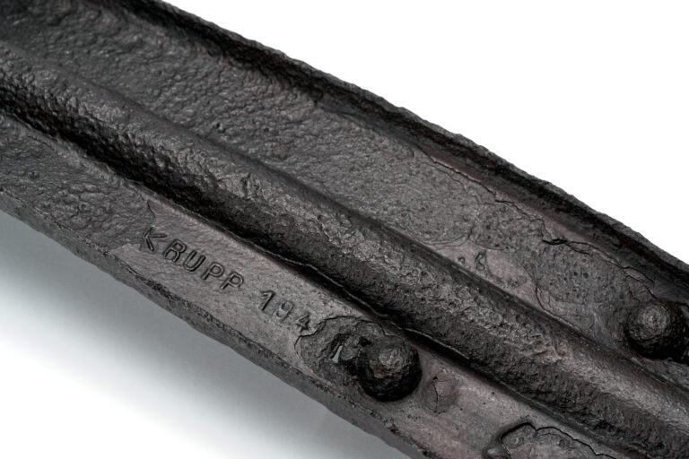 Ciężki, metalowy fragment o wydłużonym, prostokątnym kształcie, wykonany ze stali. Dłuższe boki są zagięte do góry pod kątem prostym, co tworzy kształt przypominający płytkie koryto. Pośrodku biegnie podłużne zagłębienie, a na górnej powierzchni znajdują się dwie wystające śruby. Metal jest zdeformowany i nierówny, co sugeruje działanie bardzo wysokiej temperatury lub dużego obciążenia. Na spodniej stronie widoczny jest wypukły napis z nazwą producenta Friedrich Krupp oraz fragment daty.