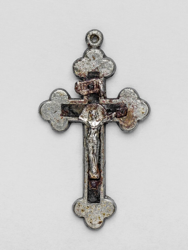Niewielki krzyżyk do noszenia na szyi. Ma formę klasycznego krzyża z końcami ramion zakończonymi trójlistnym kształtem. W środku znajduje się mała, metalowa postać ukrzyżowanego Chrystusa, z głową pochyloną w dół. Nad głową widoczna jest wąska tabliczka z napisem „INRI”. Wnętrze krzyża wypełnia ciemniejsze drewno, a metalowa powierzchnia ma miejscami przebarwienia. Jedna z rąk figurki Chrystusa jest ułamana.