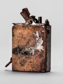 Metalowa zapalniczka benzynowa w formie małego prostopadłościanu. Na bocznej ściance ukośny napis ozdobną czcionką „Souvenier / Bruxelles”. Na górze widoczny fragment mechanizmu z blaszką i wystającymi bolcami. Powierzchnia w większości pozbawiona niklowania, z przetarciami i śladami rdzy; brak części zamknięcia.