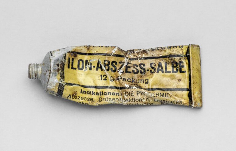 Mała, lekko zgnieciona aluminiowa tubka po maści Ilon na ropne choro. Na jasnym żółtokremowym tle widoczne czarne napisy w ramkach z nazwą preparatu i opisem zastosowań medycznych. Z jednej strony wąska, gwintowana końcówka do nakrętki.y skóry