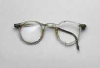 Niekompletne okulary z jasnożółtą, przezroczystą oprawką z tworzywa PVC. Zachowane prawe szkło i prawy zausznik, lewy zausznik tylko fragmentaryczny, brak lewego szkła. Oprawka o niemal okrągłych szkłach z lekko wydłużonym dołem i wygiętym mostkiem; widoczne pęknięcia i zabrudzenia.
