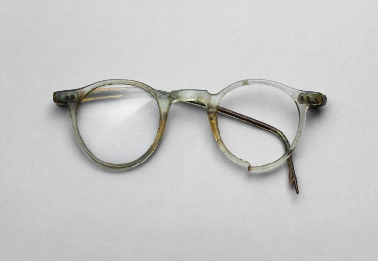Niekompletne okulary z jasnożółtą, przezroczystą oprawką z tworzywa PVC. Zachowane prawe szkło i prawy zausznik, lewy zausznik tylko fragmentaryczny, brak lewego szkła. Oprawka o niemal okrągłych szkłach z lekko wydłużonym dołem i wygiętym mostkiem; widoczne pęknięcia i zabrudzenia.