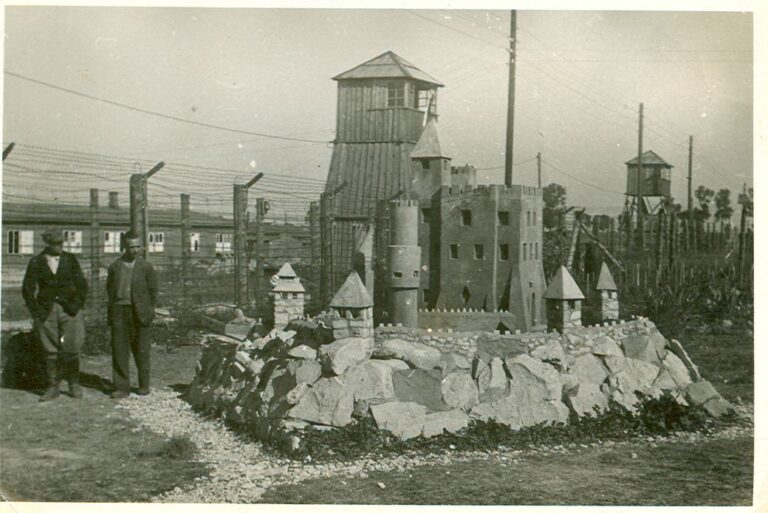 Tzw. "zameczek" - kamienna rzeźba-budowla wzniesiona przez więźniów na II polu latem 1943 r. Zdjęcie wykonane jest od wewnątrz pola w kierunku północnym. Za "zameczkiem" wieża wartownicza, a po lewej stronie zdjęcia barak nr 62. Po prawej kolejna wieża (typu "bocianie gniazdo"), w tle drzewa rosnące przy szosie piaseckiej.