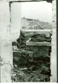 Stół sekcyjny w spalonym budynku krematorium. W pomieszczeniu widoczne rozrzucone ubrania pozostałe po ofiarach egzekucji. Za murem - ogrodzenie i baraki V pola.