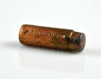 Szminka w metalowym, walcowatym opakowaniu z napisem „MIRACULUM”. Na powierzchni nasadki widoczny jest dekoracyjny wzór z pionowych linii oraz nazwa firmy i stary numer inwentarzowy „B/49/99”. W dolnej części oprawki znajduje się niewielkie owalne zakończenie zabezpieczające nasadkę przed zsunięciem. Opakowanie nosi ślady zużycia i drobnych uszkodzeń, w tym mały ubytek na górnej powierzchni nasadki.